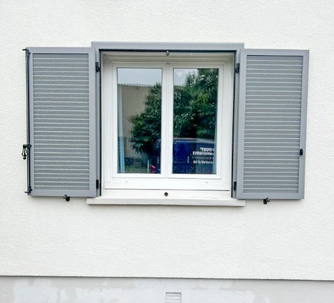 Façade extérieure d'habitation avec fenêtre à deux vantaux ouvrants, équipée de volets battants en aluminium gris à lames orientables