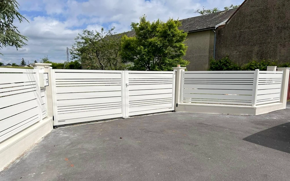 Portail coulissant moderne en aluminium anthracite avec panneaux ajourés et grillage métallique central, installé entre piliers maçonnés blancs sur entrée gravillonnée d'une propriété résidentielle